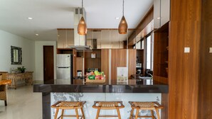 A modern kitchen with a central island, wooden bar stools, pendant lighting, and a dining area with a wicker table and chairs. - Magenta Villa 6 BR With Pool Movieroom Billiard (Bandung)