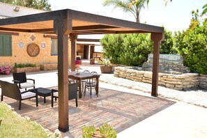 An outdoor patio with a wooden pergola, stone flooring, and a seating area with chairs and a table.