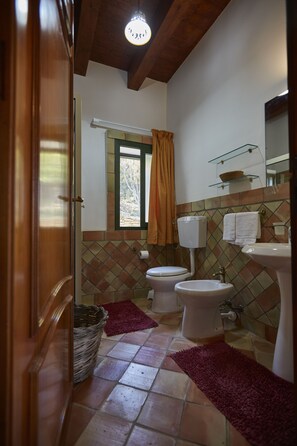 A bathroom with a toilet, bidet, and sink, wooden ceiling beams, a window with a curtain, and a tiled floor.