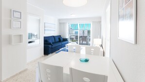 A modern hotel room with a white color scheme, a blue sofa, a dining table with chairs, and a balcony with a striped awning. - Beautiful condo family near to the beach. Pool / Wifi - only families (Playa de Gandía)