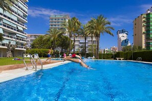 Une piscine où une personne plonge, entourée de palmiers et de bâtiments modernes.
