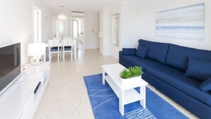 A modern living room with a blue sofa, a white coffee table with a plant, a TV, and a dining area with white chairs and a table. - Beautiful condo family near to the beach. Pool / Wifi - only families (Playa de Gandía)