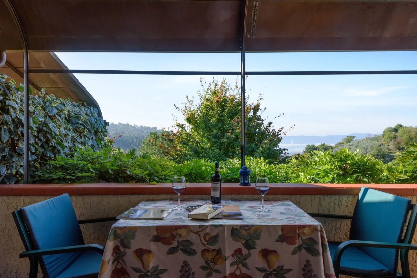 Une table d’extérieur recouverte d’une nappe à motifs floraux, deux chaises, une bouteille de vin et un verre de vin.