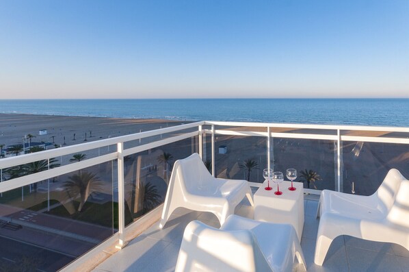 A balcony with white plastic chairs and tables, overlooking a beach and the ocean.