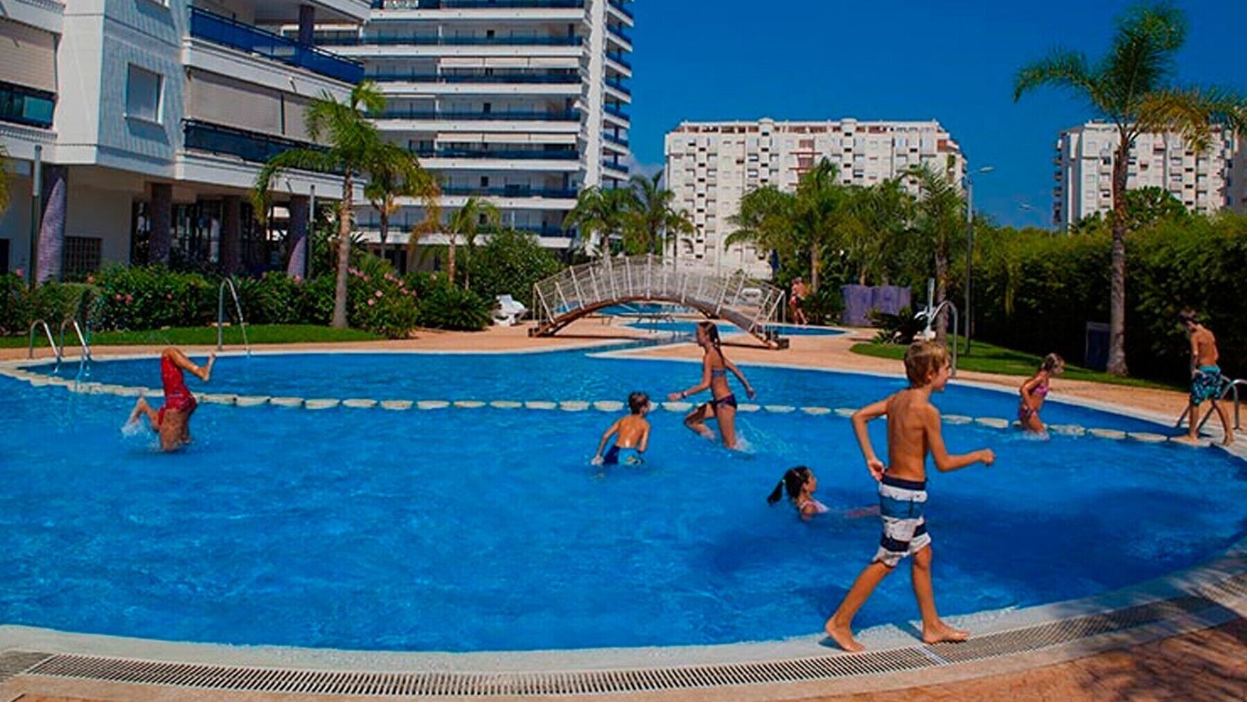 Une piscine où des gens nagent et s’amusent, entourée de palmiers et d’immeubles modernes.