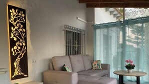 A modern living room with a beige sectional sofa, a decorative wall piece, a small side table with a potted plant, and a view of the outdoors through a large window. - Modern villa with large garden and close to the sea (Catral)