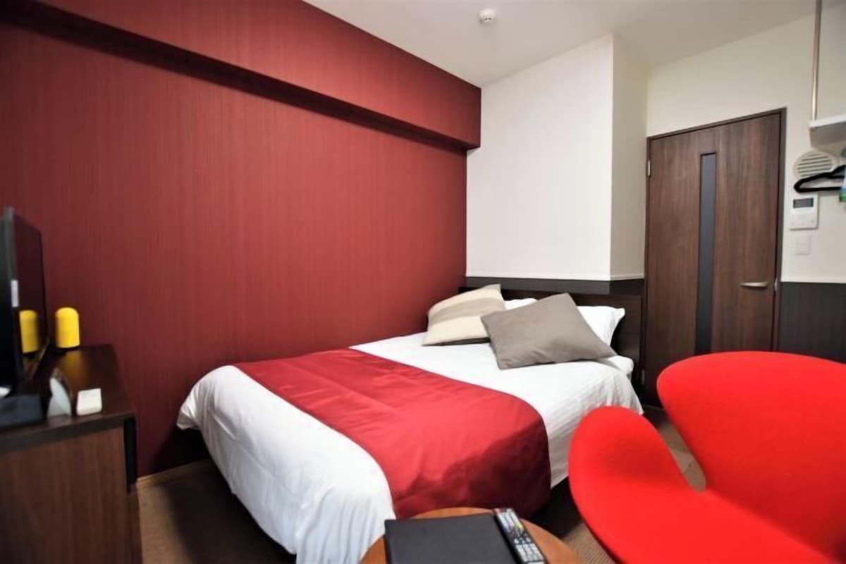 Habitación de hotel con cama, una silla roja, un escritorio con lámpara y un armario.