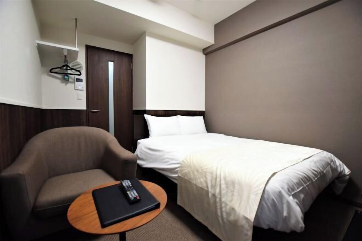 Habitación de hotel con cama, una silla, una mesita y perchero.