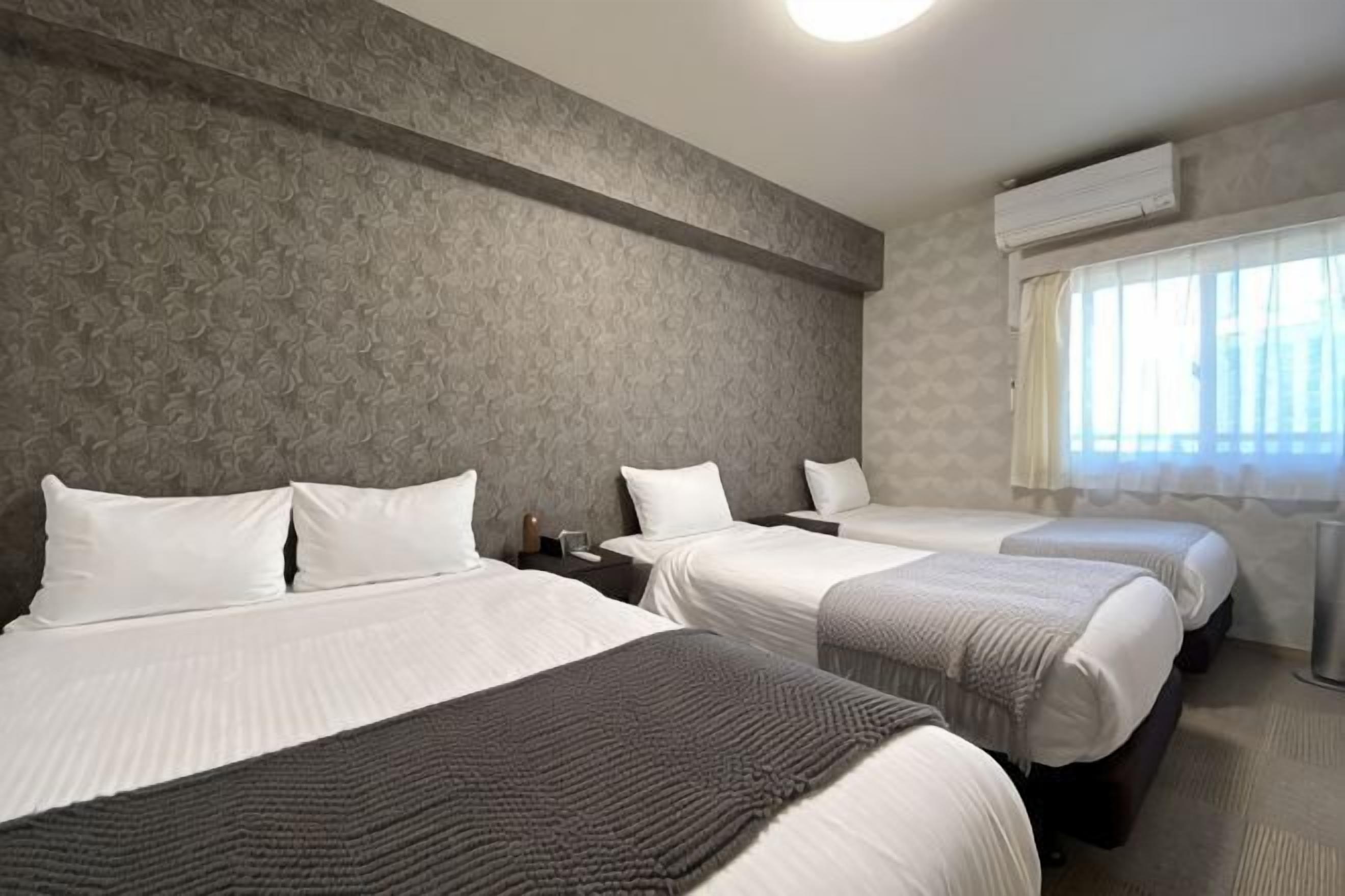 Habitación de hotel con tres camas, un ventanal con cortinas y un aire acondicionado instalado en la pared.
