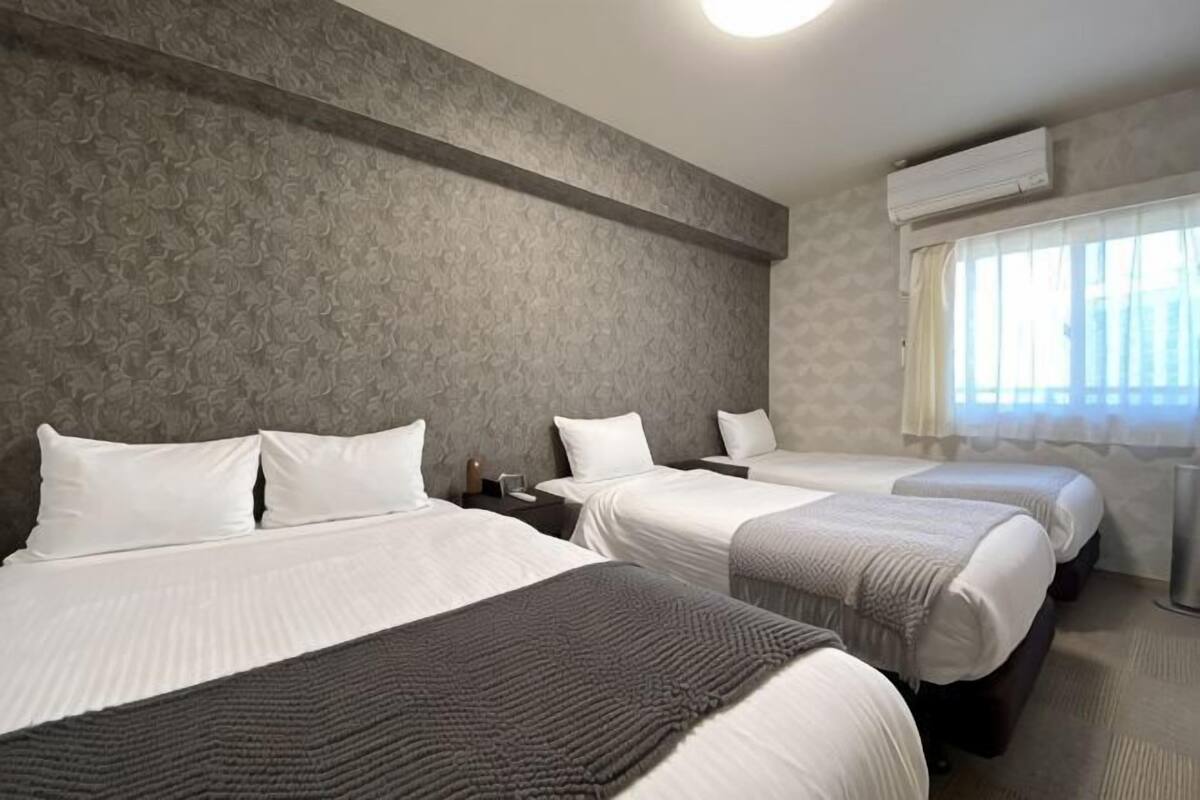 Habitación de hotel con tres camas, un ventanal con cortinas y un aire acondicionado instalado en la pared.