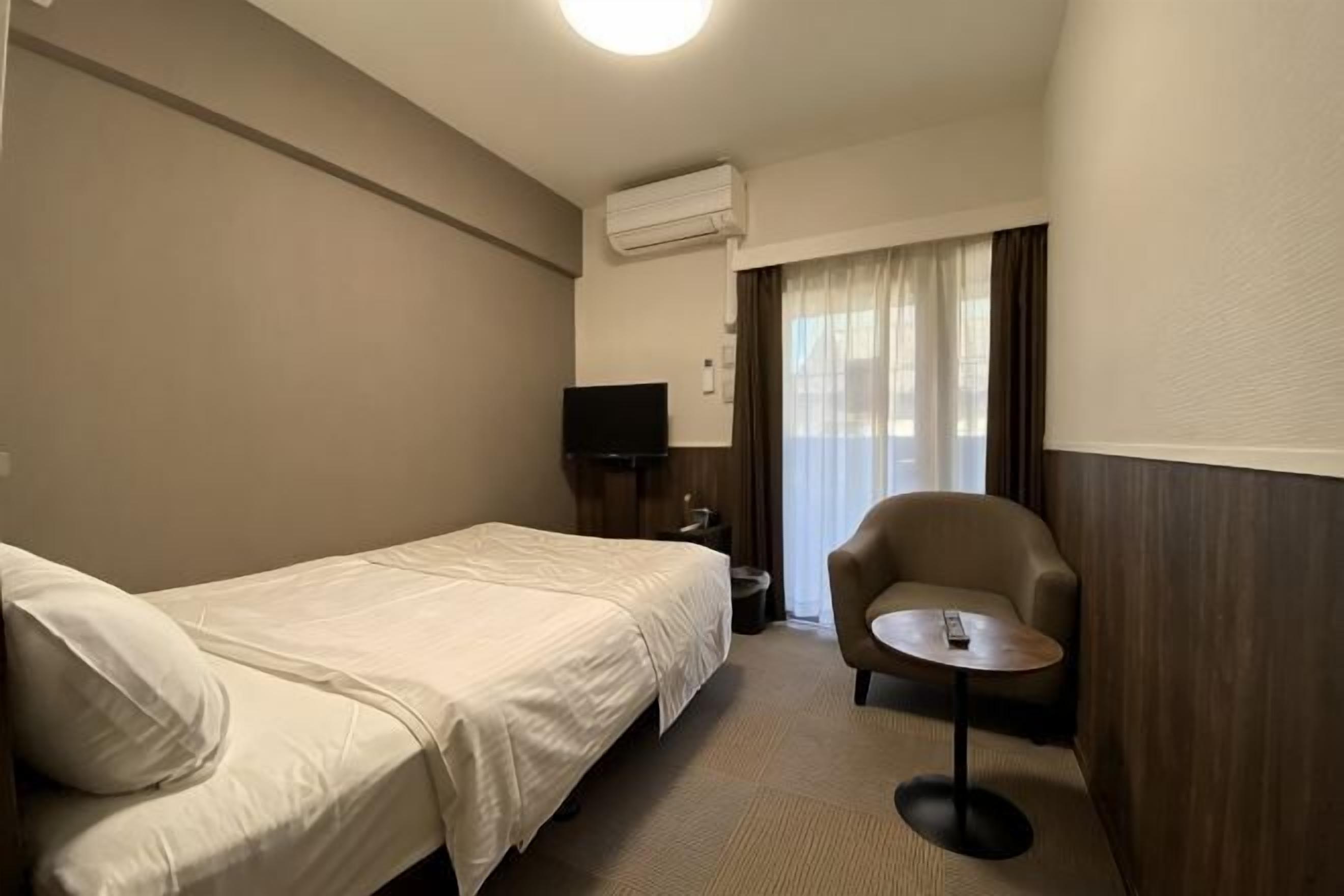 Habitación de hotel con una cama individual, una silla, una mesita pequeña, un televisor y un equipo de aire acondicionado.