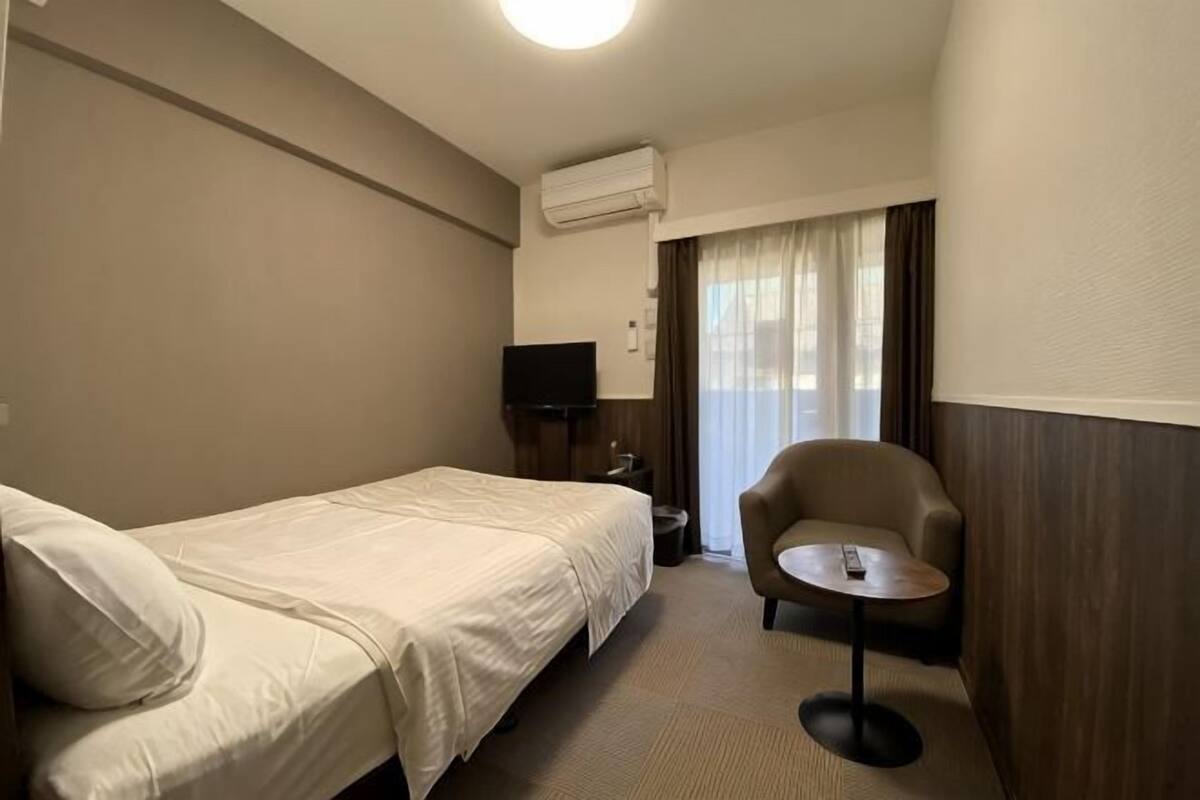 Habitación de hotel con una cama individual, una silla, una mesita pequeña, un televisor y un equipo de aire acondicionado.