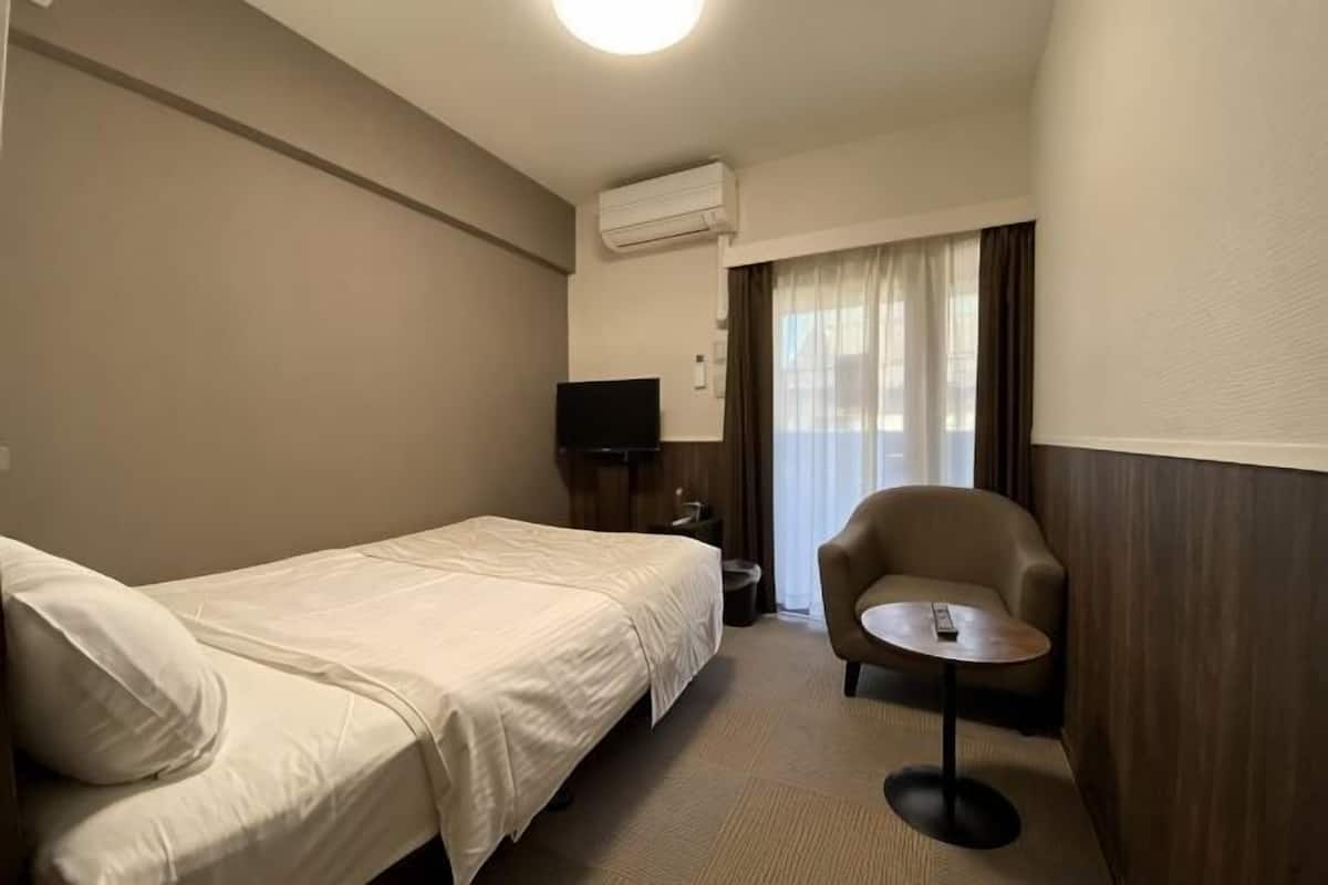 A hotel room with a single bed, a chair, a small table, a TV, and an air conditioning unit.