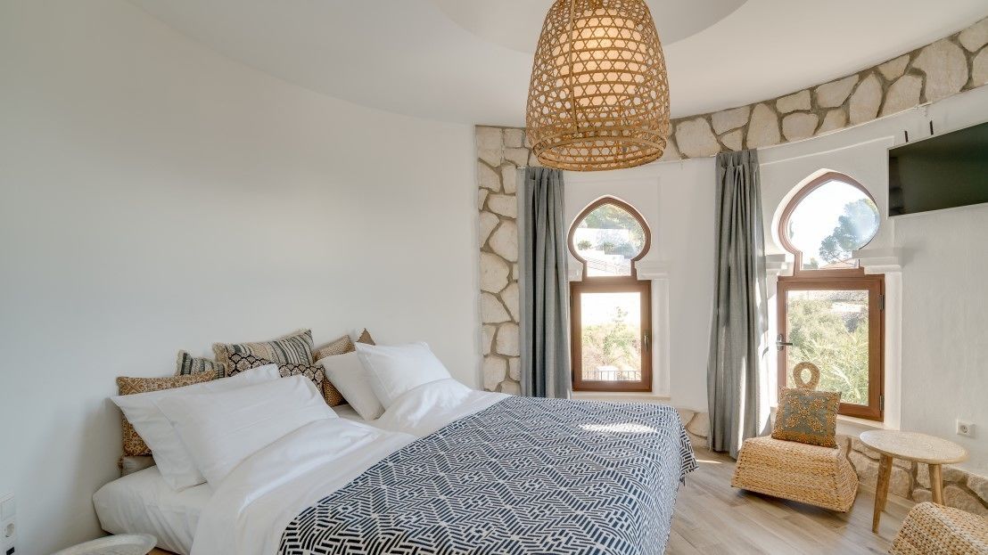 Une chambre avec un grand lit, un luminaire suspendu, des murs en pierre et deux fenêtres avec des rideaux.