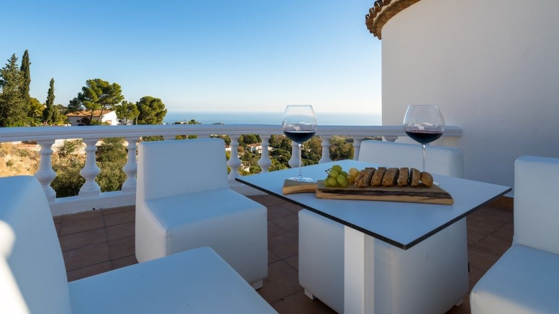 Een terras met witte meubels, een tafel met een fruitschaal en wijnglazen, en uitzicht op de zee.