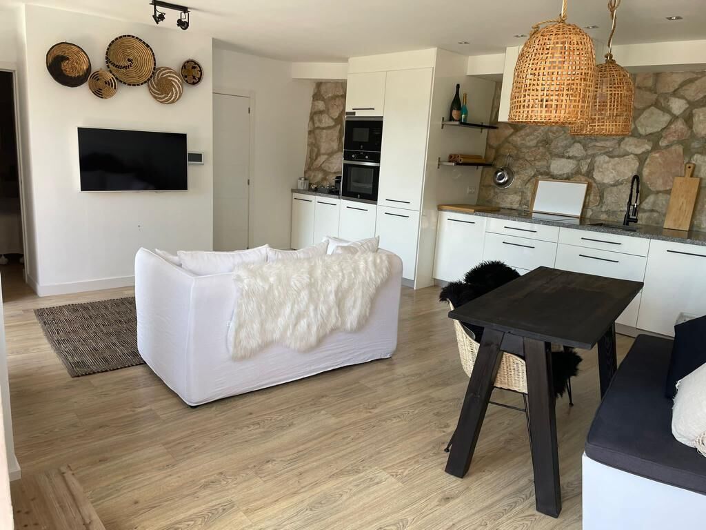 Ein modernes Wohnzimmer mit einem weißen Sofa, einem schwarzen Esstisch und einer Küchenzeile mit weißen Schränken und einer Arbeitsplatte aus Stein.