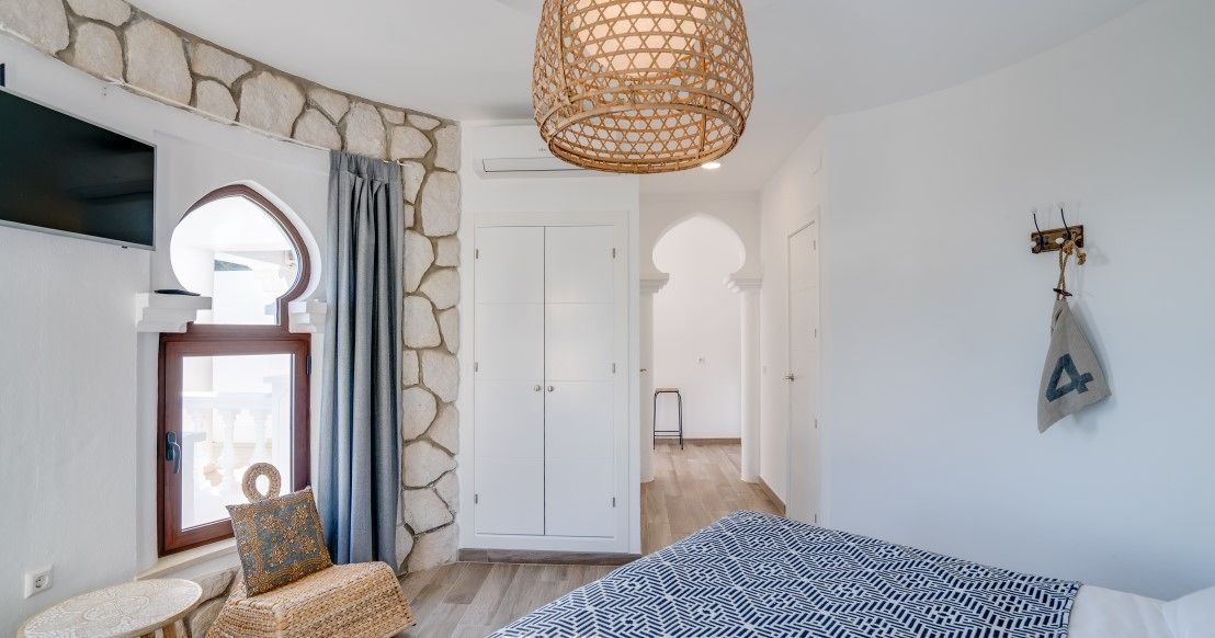 A bedroom with a stone wall, a bed with a patterned bedspread, a wicker chair, a hanging light fixture, and a wardrobe.