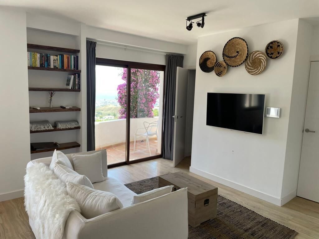 Un salon moderne avec un canapé blanc, un téléviseur à écran plat fixé au mur, une bibliothèque et un balcon offrant une vue dégagée.