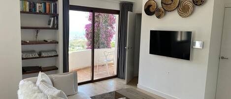 Un salon moderne avec un canapé blanc, un téléviseur à écran plat fixé au mur, une bibliothèque et un balcon offrant une vue dégagée.