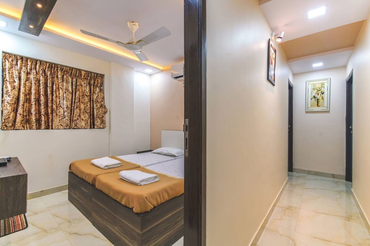 A hotel room with a bed, a desk, a ceiling fan, and a hallway with a door and a picture on the wall.
