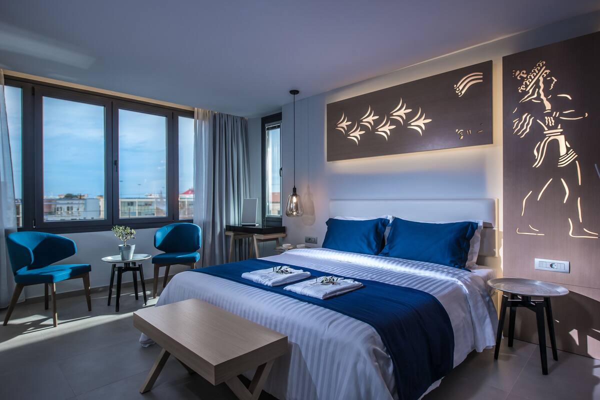 Ein modernes Hotelzimmer mit einem großen Bett, zwei blauen Stühlen, einem kleinen Tisch und Blick auf die Stadt durch große Fenster.