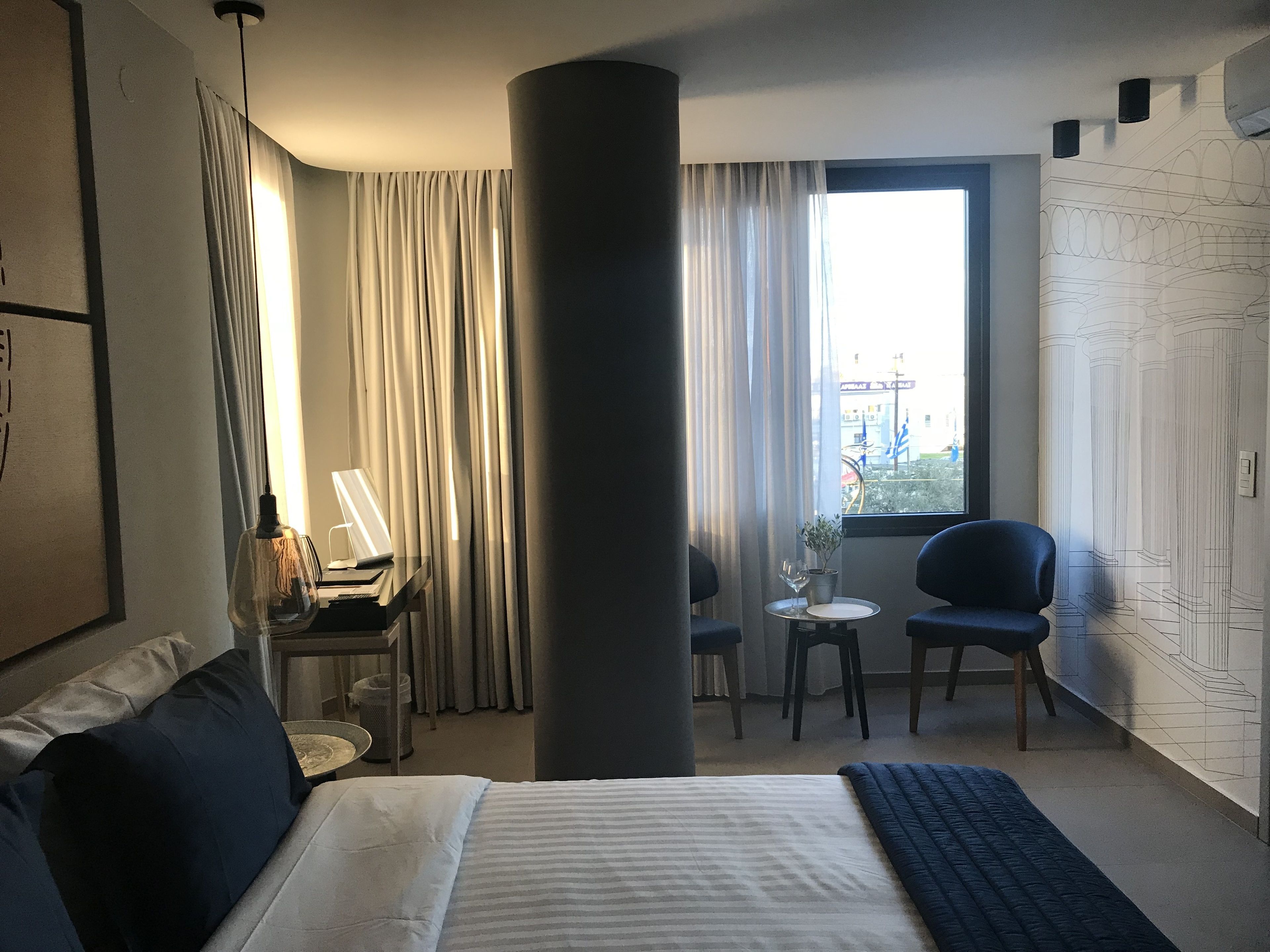 A hotel room with a bed, a desk with a lamp, a chair, a small table with a plant, and a window with curtains.