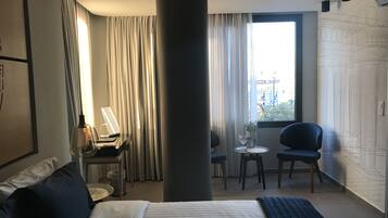 A hotel room with a bed, a desk with a lamp, a chair, a small table with a plant, and a window with curtains.