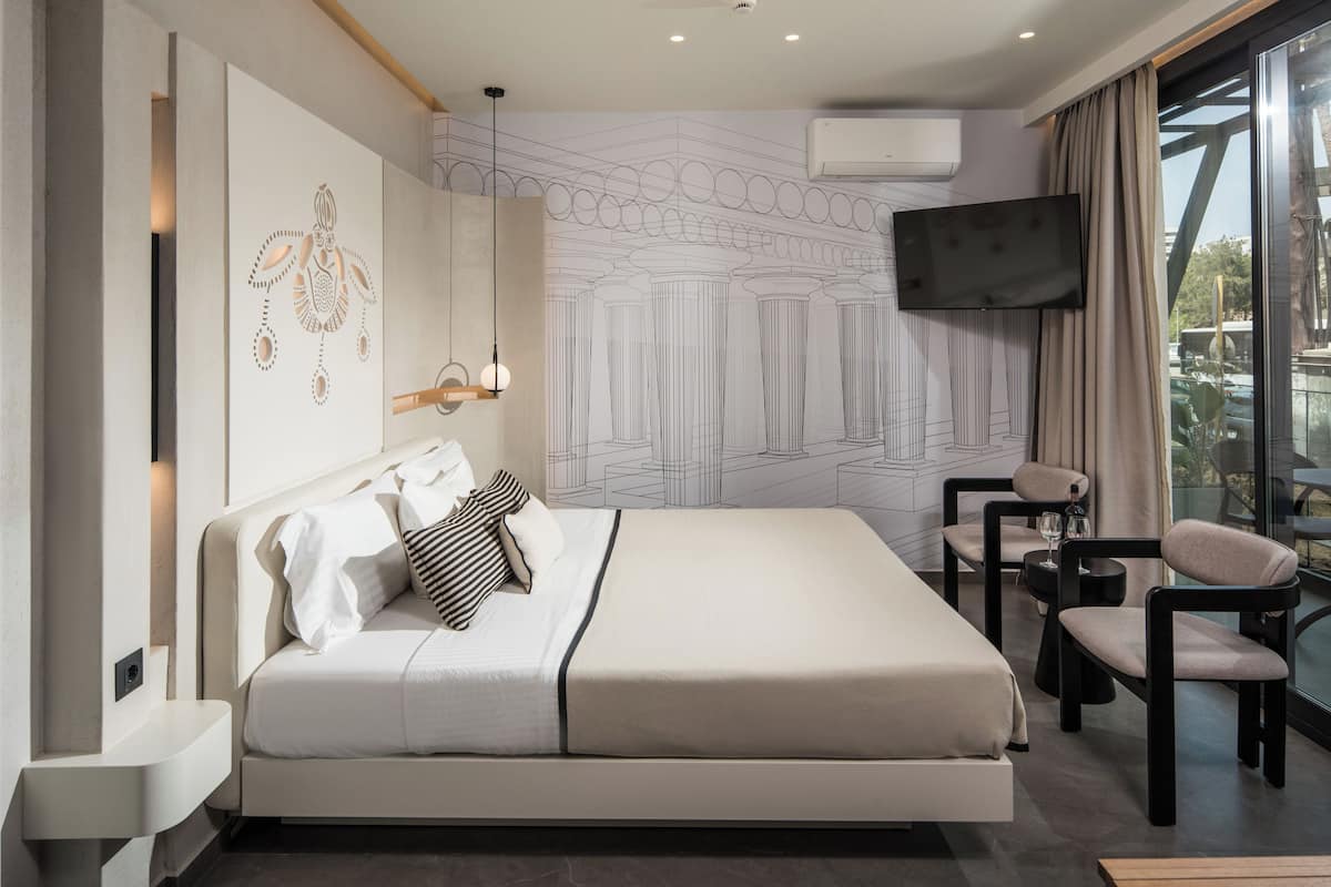 Ein modernes Hotelzimmer mit einem großen Bett, einer Sitzecke mit Tisch und Stühlen, einem an der Wand montierten Flachbildfernseher und einem großen Fenster mit Blick ins Freie.