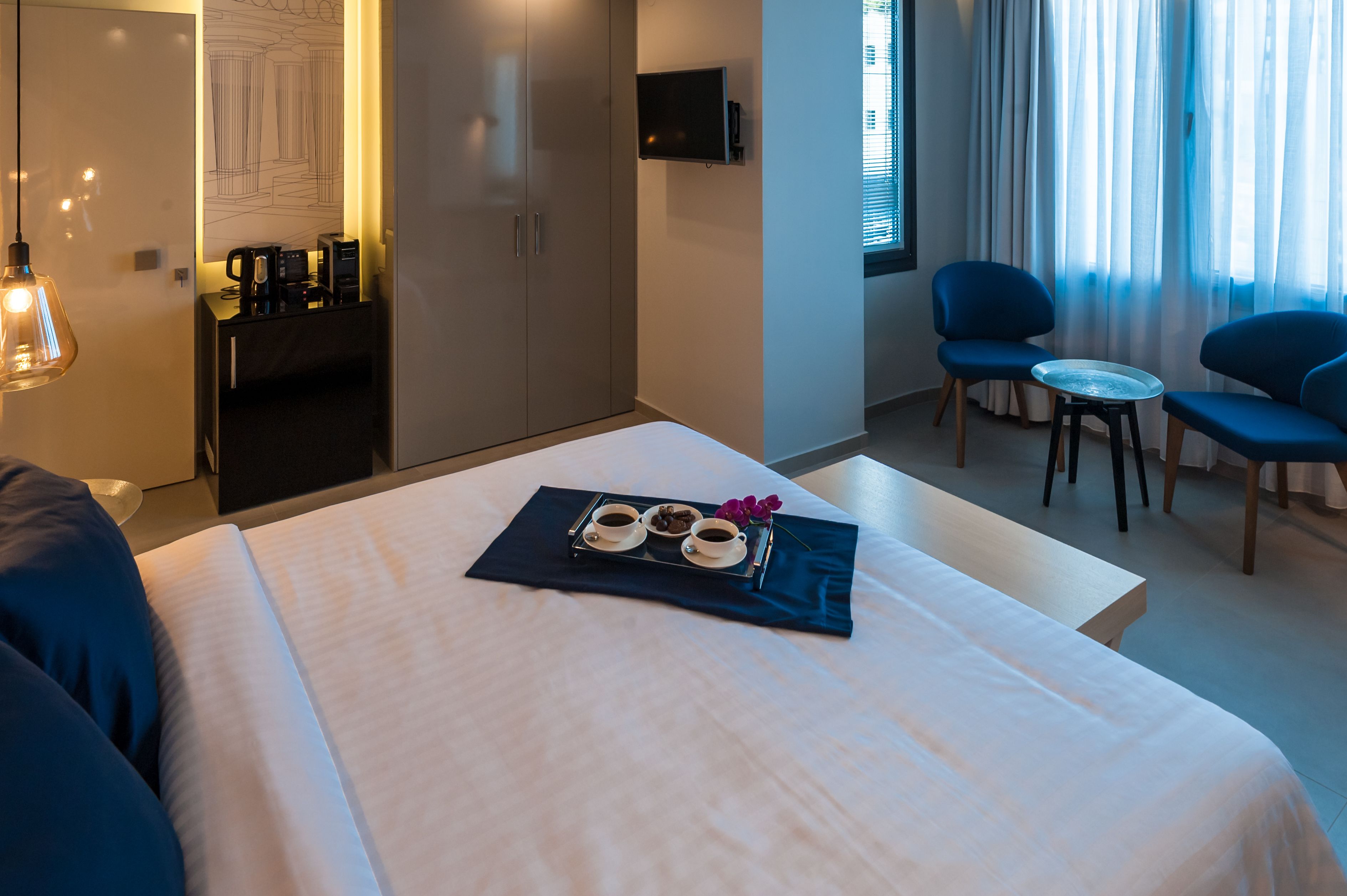 Ett hotellrum med en säng, ett litet bord med ett bricka med kaffe och bakverk, två stolar, en tv på väggen och ett fönster med gardiner.