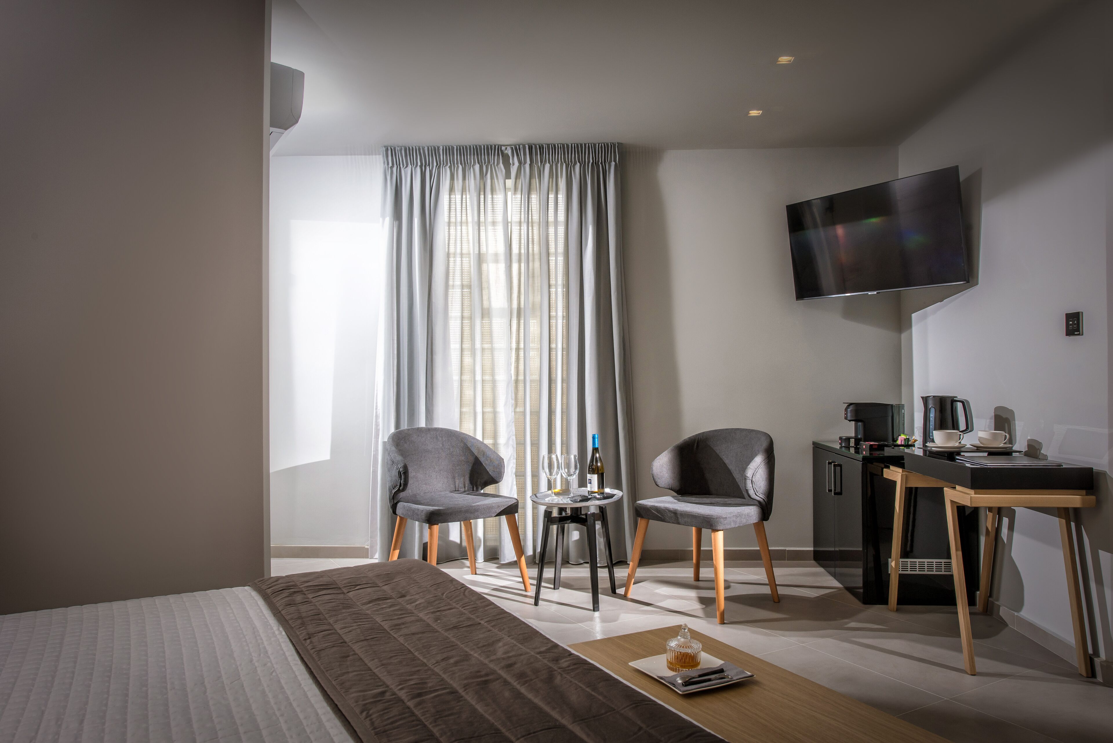 Ein modernes Hotelzimmer mit einem Bett, zwei Stühlen, einem kleinen Tisch mit einer Flasche, Gläsern und einer Tasse, einem Schreibtisch mit einer Kaffeemaschine und einem an der Wand befestigten Fernseher.