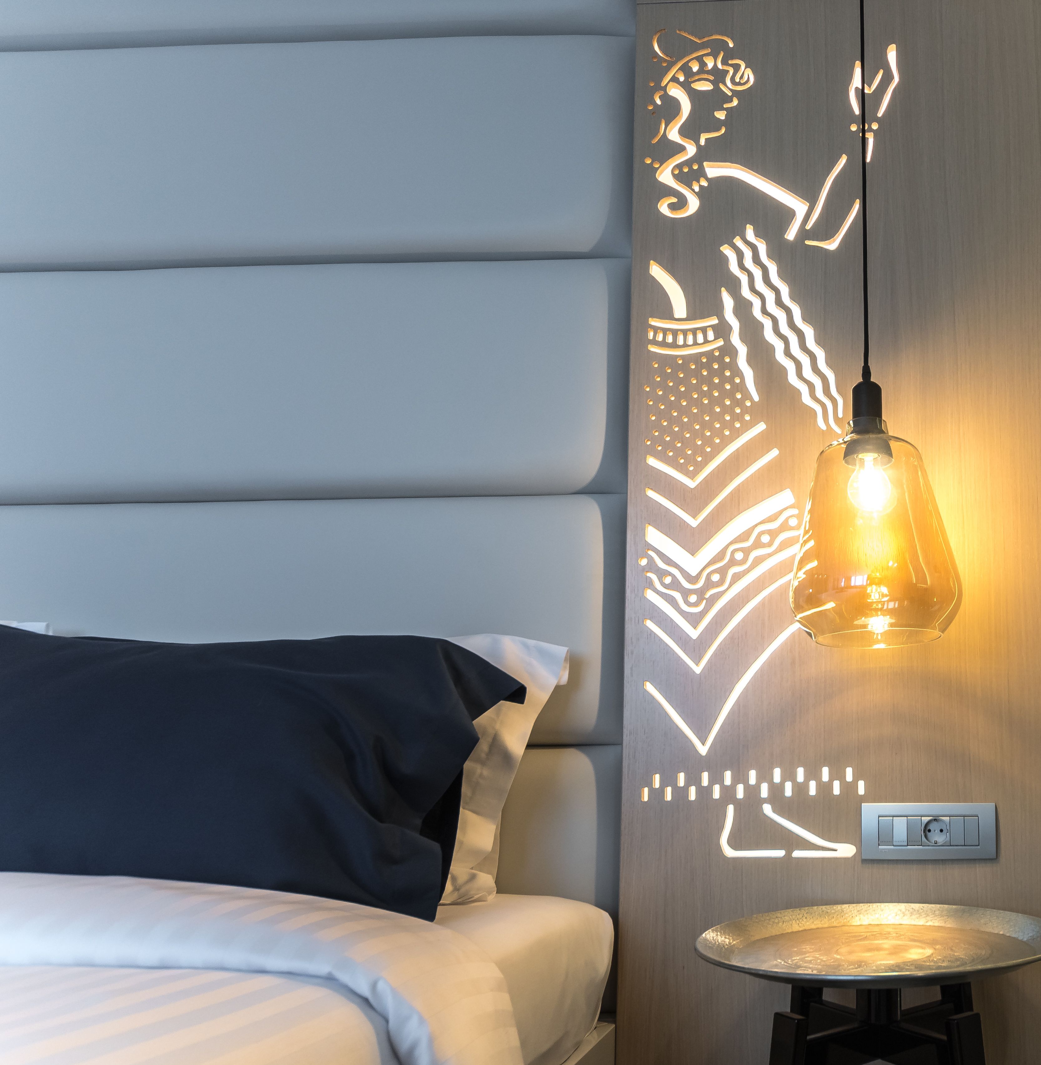A modern hotel room with a bed, a wall-mounted light fixture, and a small table.