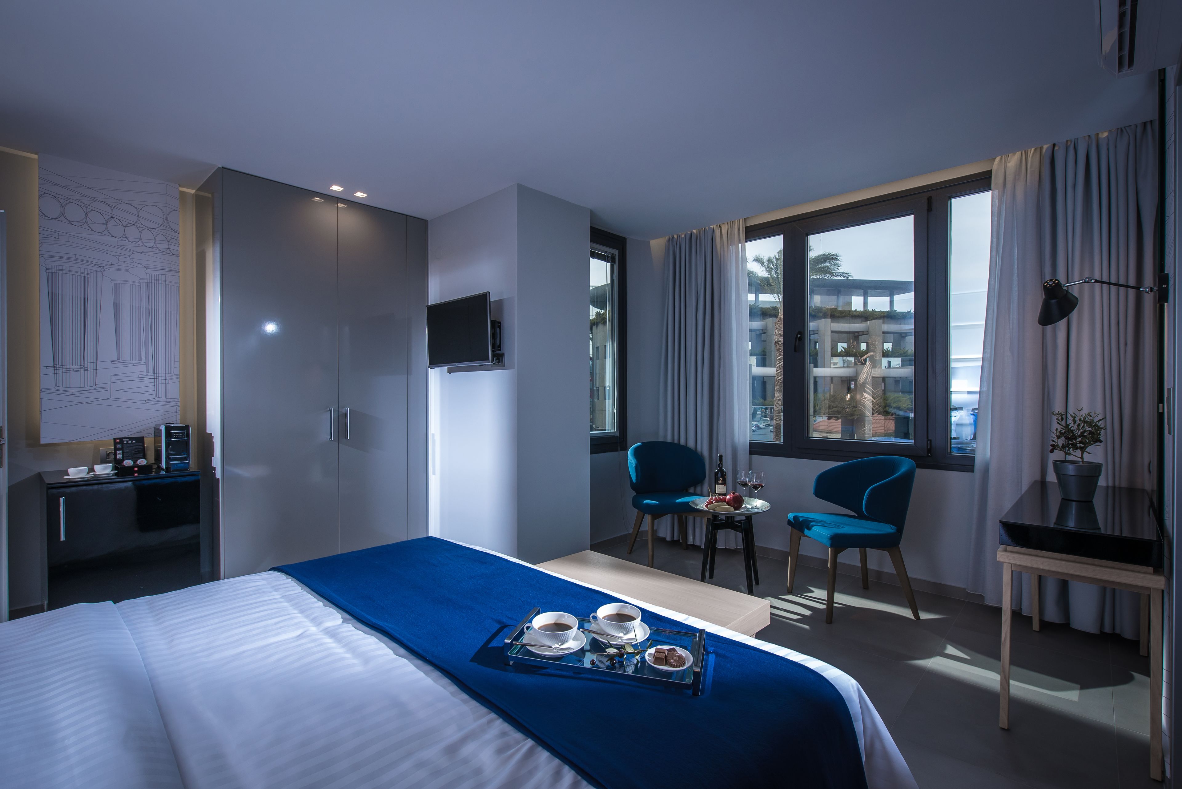 A hotel room with a bed, a desk, a TV, a dining area with chairs, and a window with curtains.