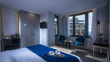 A hotel room with a bed, a desk, a TV, a dining area with chairs, and a window with curtains.