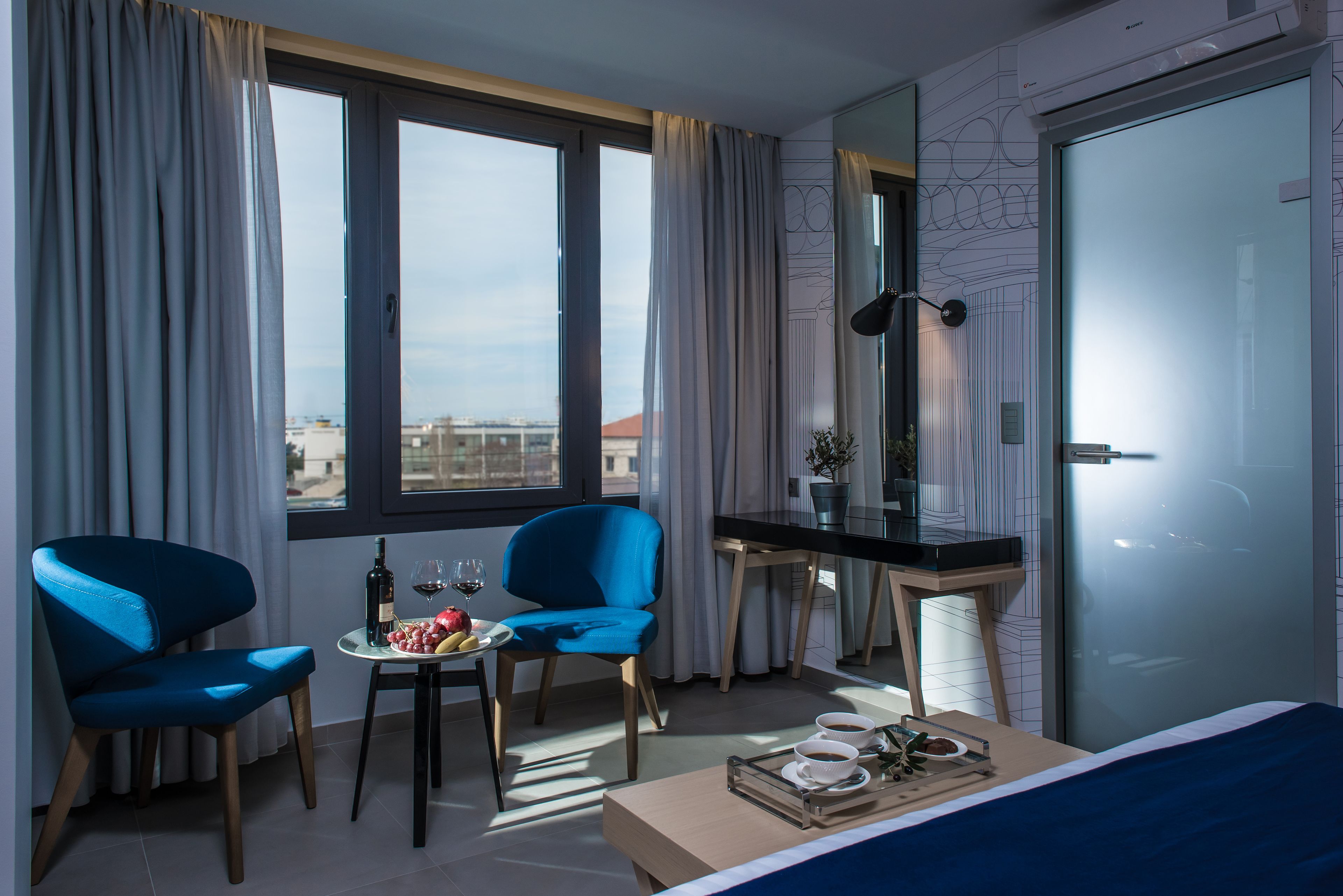 Une chambre d’hôtel moderne avec une grande fenêtre, deux fauteuils bleus, une petite table sur laquelle se trouve un plateau avec de la nourriture et des boissons, un bureau et un lit.