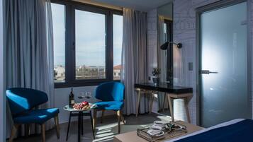 Une chambre d’hôtel moderne avec une grande fenêtre, deux fauteuils bleus, une petite table sur laquelle se trouve un plateau avec de la nourriture et des boissons, un bureau et un lit.