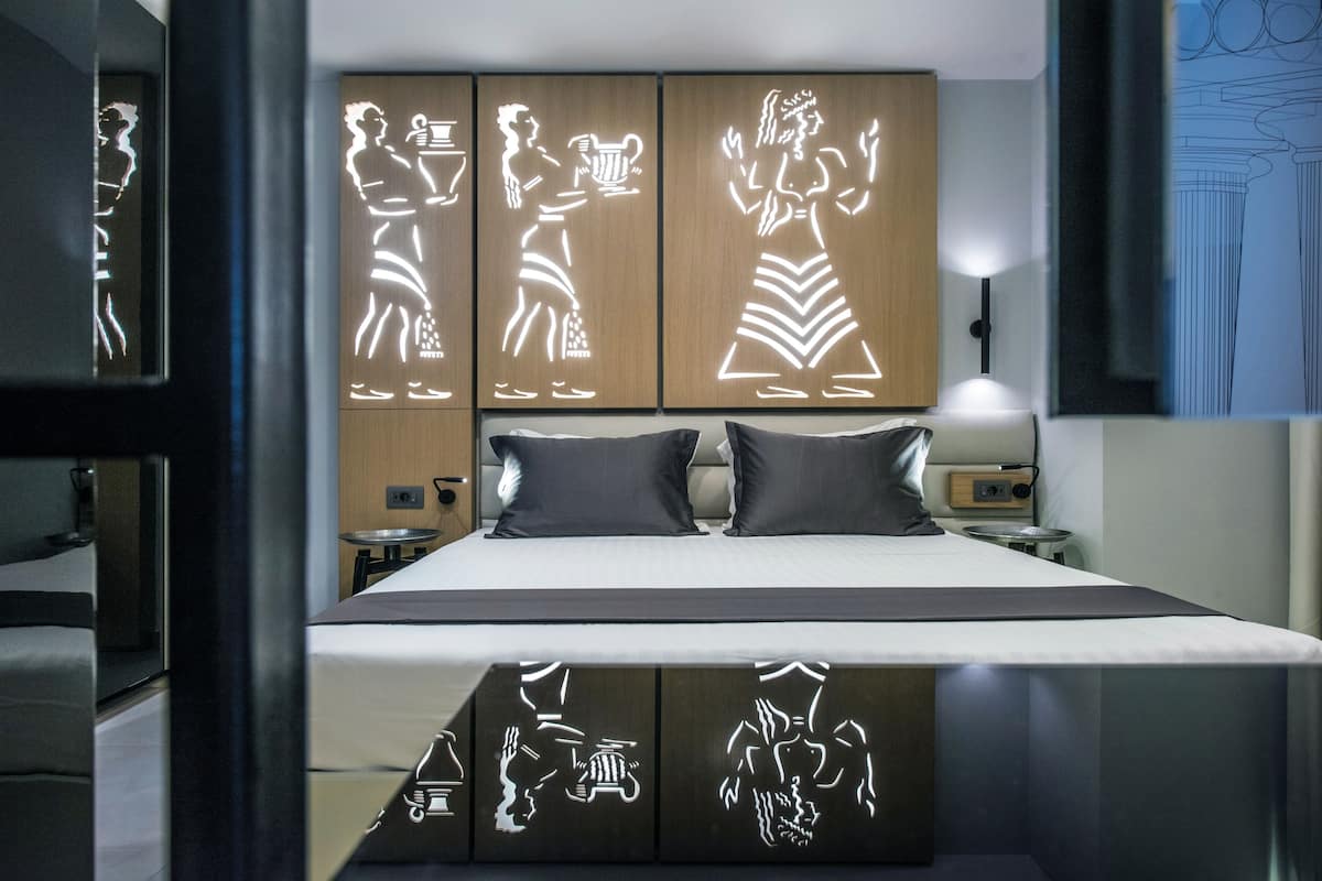 Ein modernes Hotelzimmer mit einem großen Bett, einem hölzernen Kopfteil mit beleuchteter Kunst, einem Nachttisch mit Lampe und einem zusätzlichen Beistelltisch.