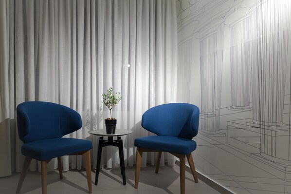 A room with two blue chairs, a small table with a potted plant, and a large window with vertical blinds. - LAVRIS City Suites (Heraklion)