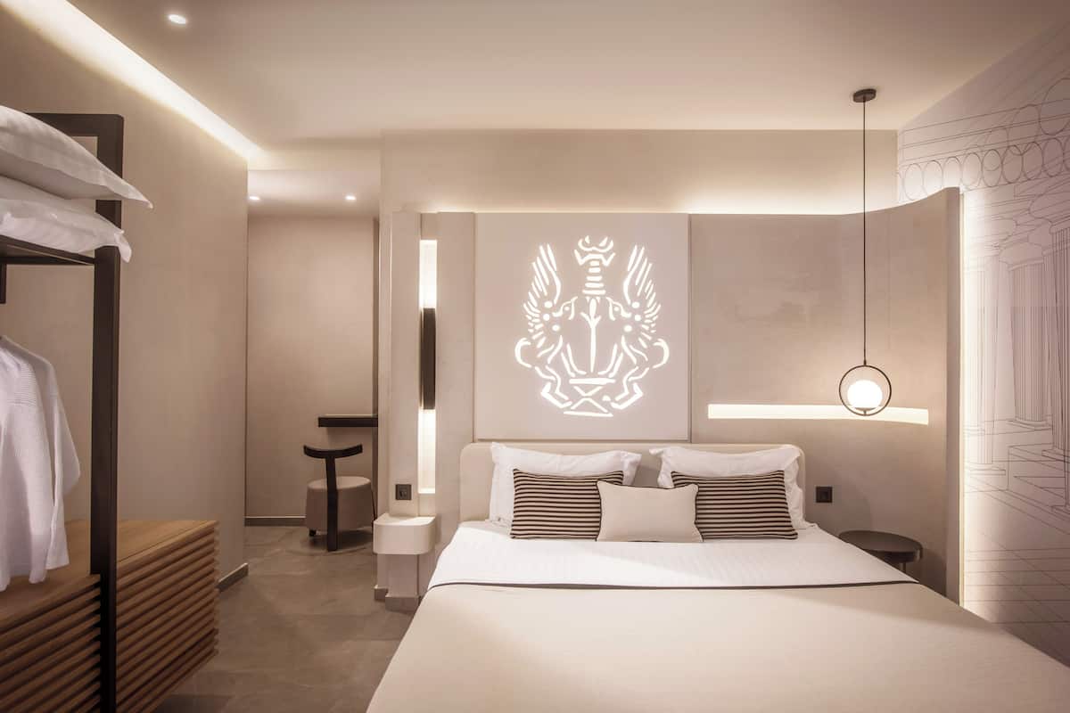 Ein modernes Hotelzimmer mit einem ordentlich bezogenen Bett, einem Nachttisch, einer Wandleuchte und einem großen Emblem an der Wand.