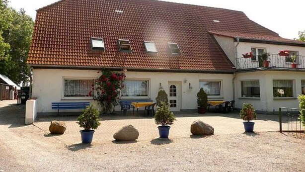 A building with a red-tiled roof, multiple windows, and a paved courtyard with potted plants and a bench. - Room 1 - Rügen apartment 52 (Schaprode auf Rügen)