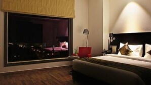 Une chambre d’hôtel moderne avec une grande fenêtre, un lit avec du linge de lit blanc, une chaise rouge et une table de chevet avec une lampe.