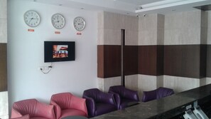 A waiting area with three clocks indicating different time zones, a flat-screen TV, and a reception desk with a counter. - Joy Inn & Suites (Tijara)