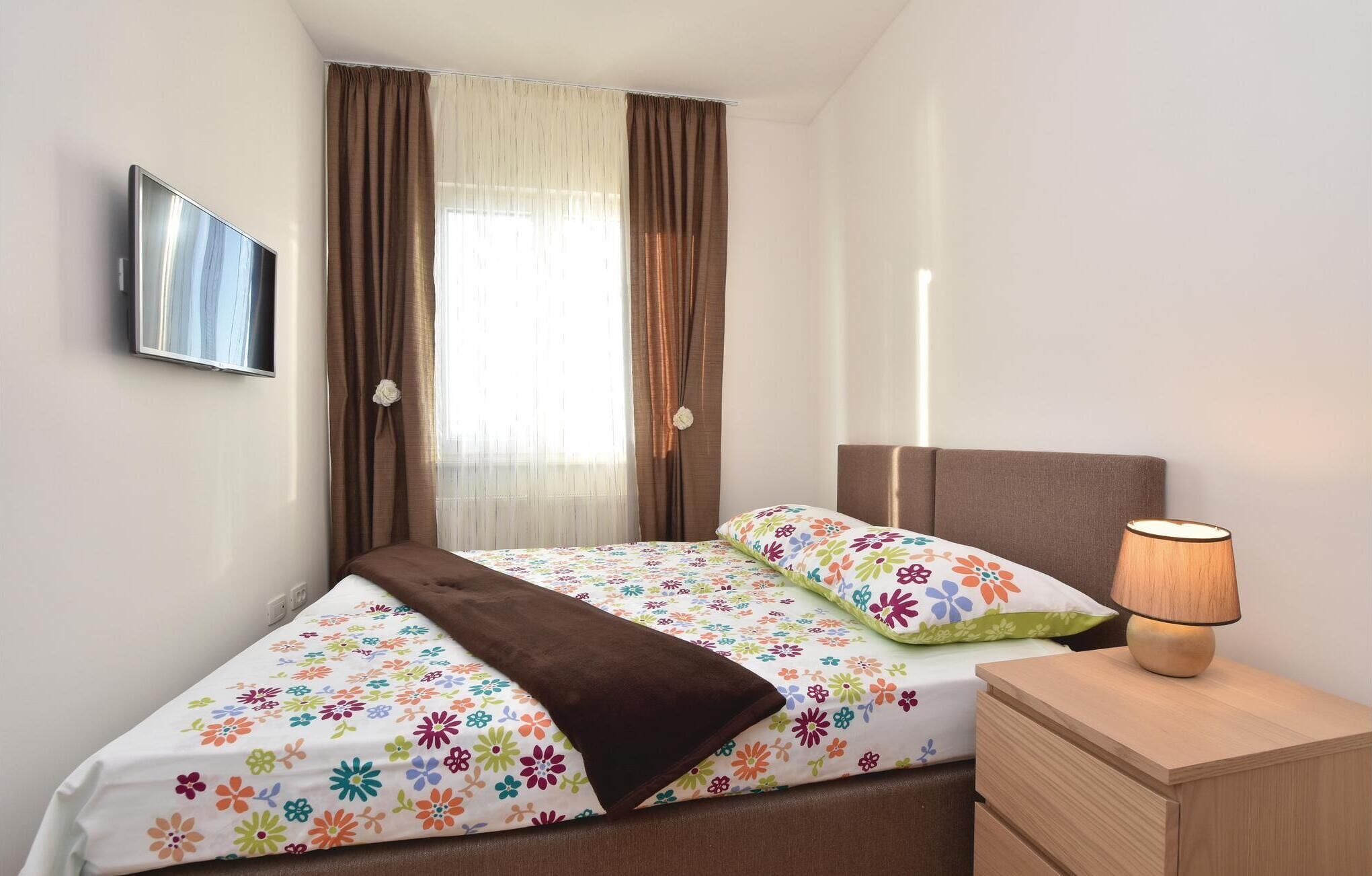 Un dormitorio ordenado con una cama, una lámpara de noche, un televisor montado en la pared y una ventana con cortinas.