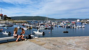A marina with boats docked, a pier with people sitting, and a hilly landscape in the background. - Lobeira do Mar (O Pindo-Carnota, LA Coruña)