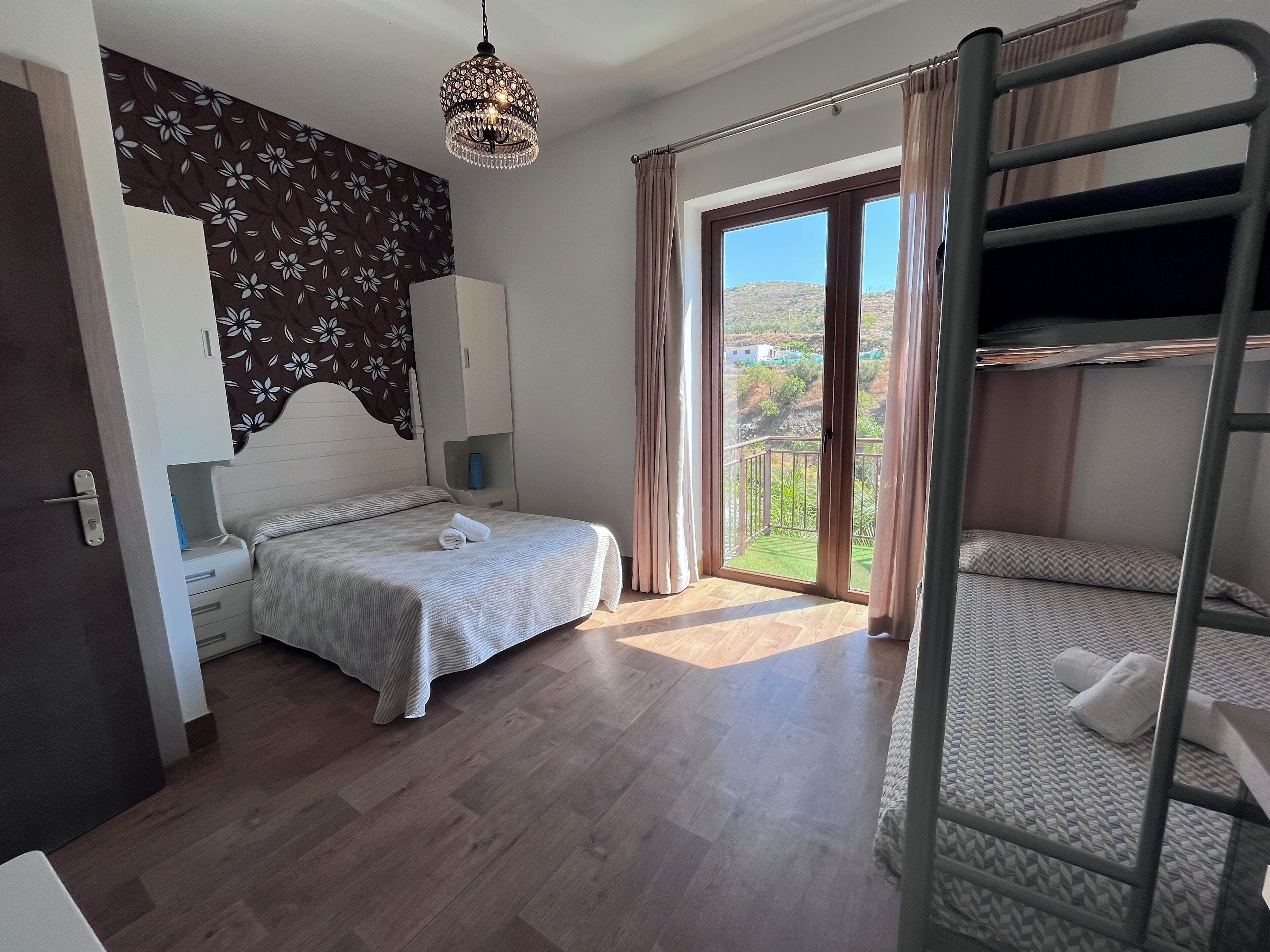 A bedroom with a bunk bed, a single bed, a wardrobe, a bedside table, a hanging light fixture, and a view of the outdoors.