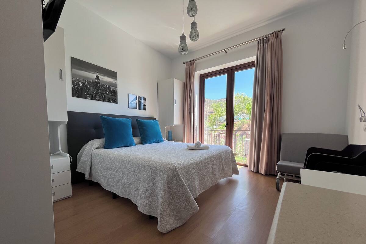 Un dormitorio con cama, escritorio y balcón con cortinas.