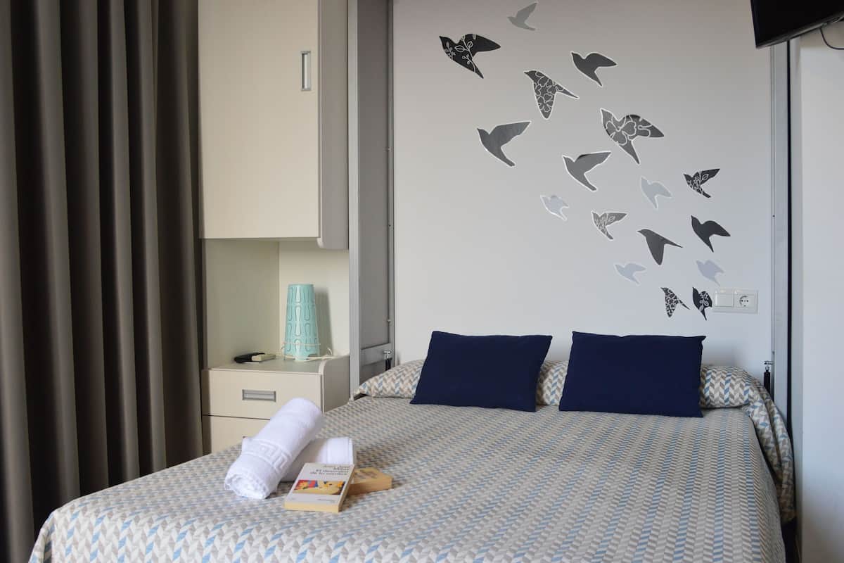 Una cama bien hecha con ropa de cama estampada, una pared con diseños de pájaros, una mesita de noche con una lámpara y una ventana con cortinas.