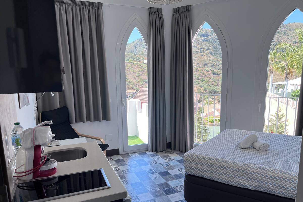 Una habitación de hotel con cama, escritorio con lavamanos y ventanas amplias con cortinas.