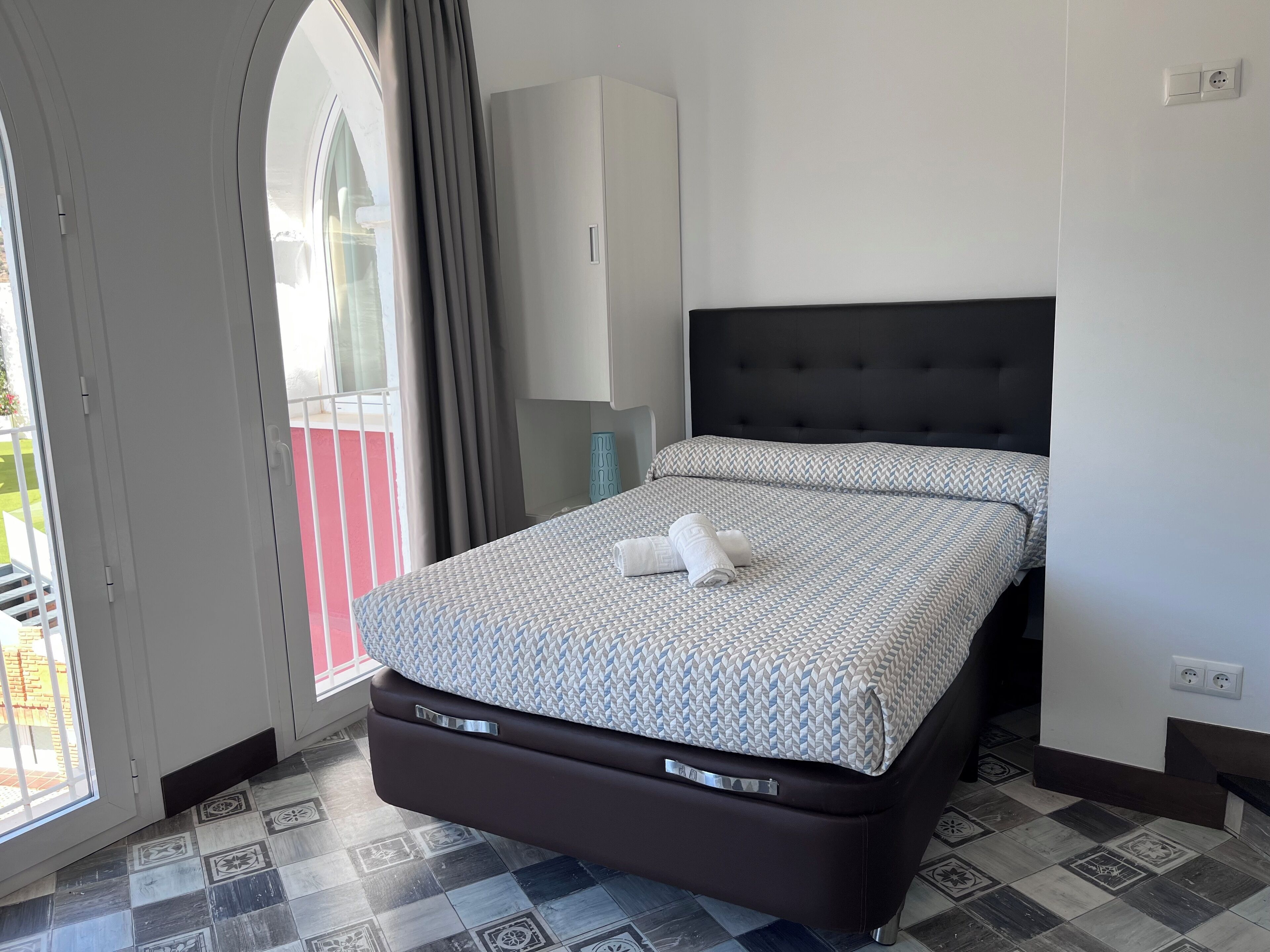 A bedroom with a bed, a window with curtains, a wardrobe, and a tiled floor.