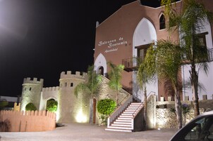 Un edificio con fachada similar a un castillo, llamado "Balcones de Bentomiz Apartahotel", con paredes de piedra, ventanas en arco y una escalera que conduce a un nivel superior.