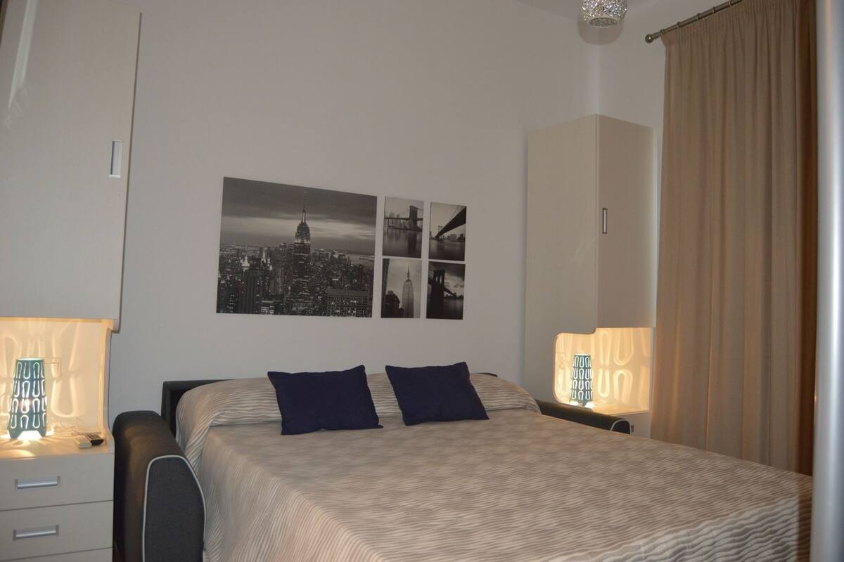 Una habitación de hotel con una cama, dos almohadas, una mesita de noche, una lámpara y una pared con tres fotografías en blanco y negro.